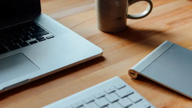 A laptop, keyboard, notebook, and coffee mug on a wooden desk.