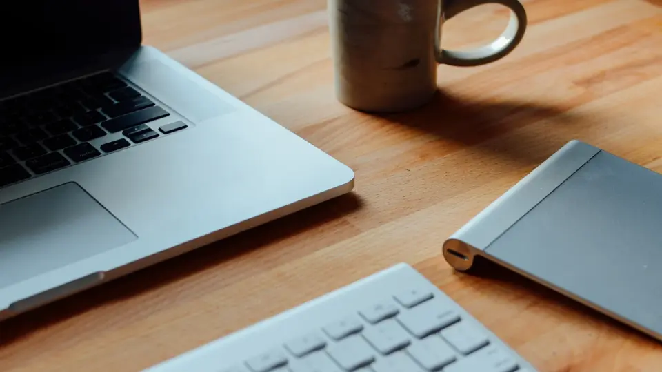 A laptop, keyboard, notebook, and coffee mug on a wooden desk.