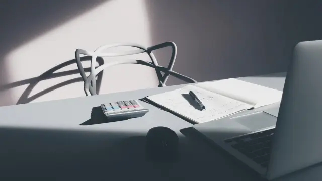 A calculator, open notebook, pen, and laptop on a desk in window light.