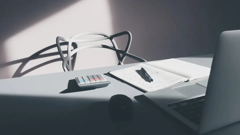 A calculator, open notebook, pen, and laptop on a desk in window light.